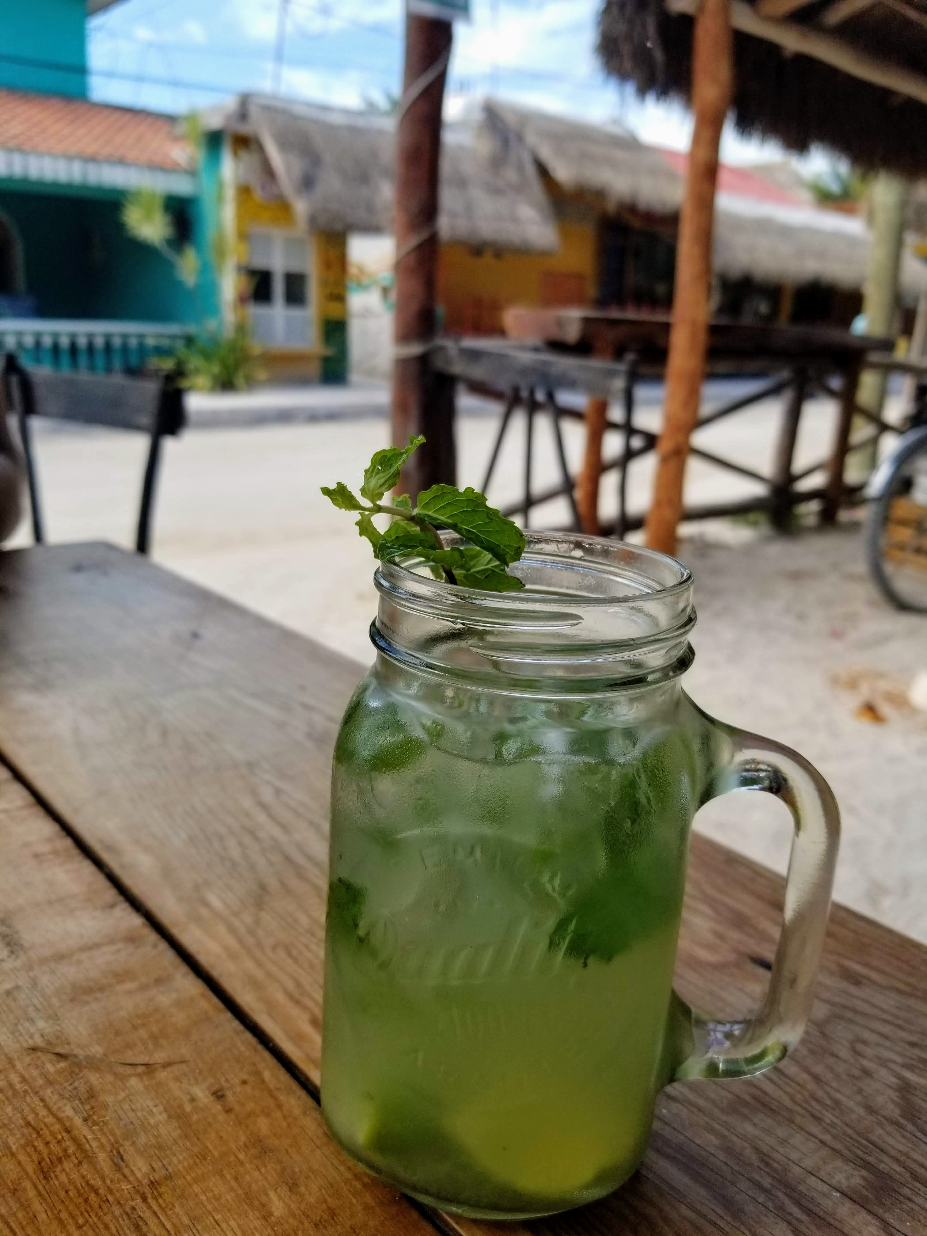A mojito in a mason jar glass sits atop a dark brown plank table on the patio of a restaurant.