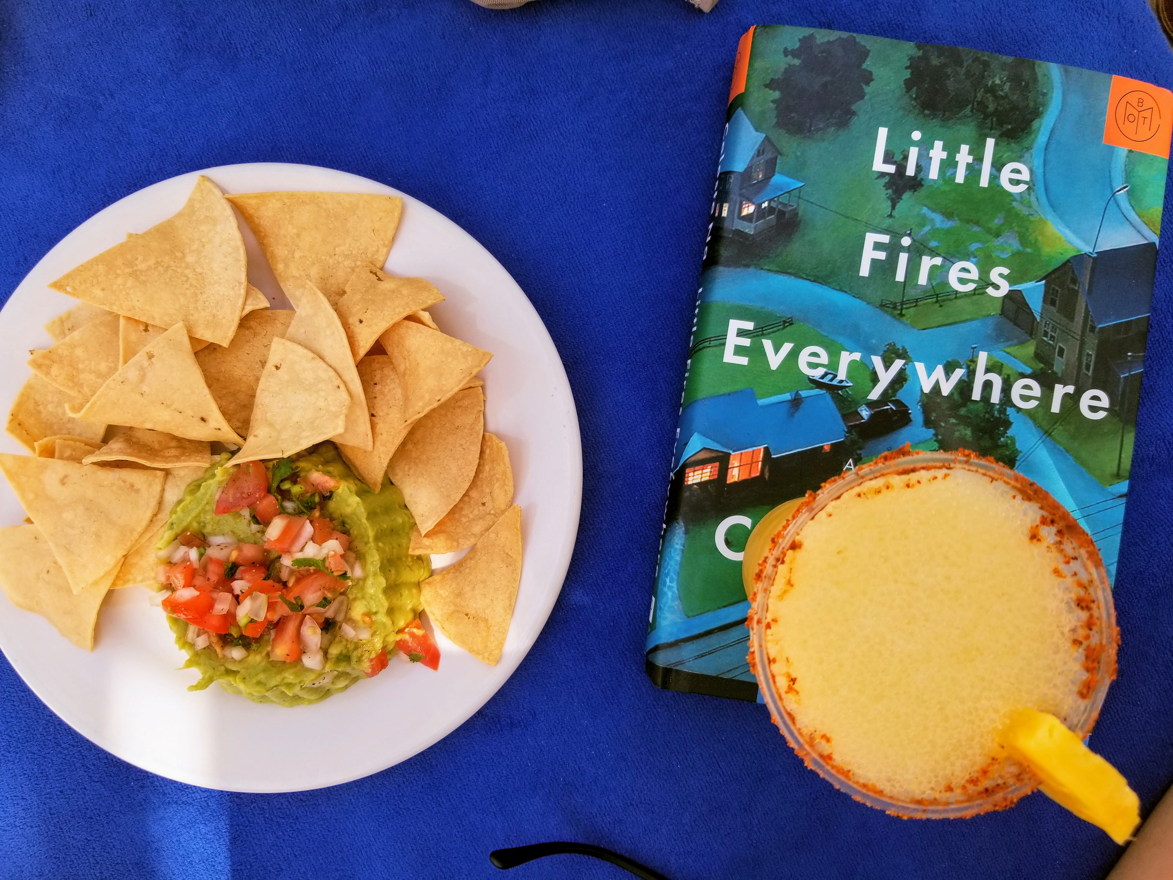 A plate of guacamole and chips is to the left of the book Little Fires Everywhere. On top of the book is a pineapple margarita.