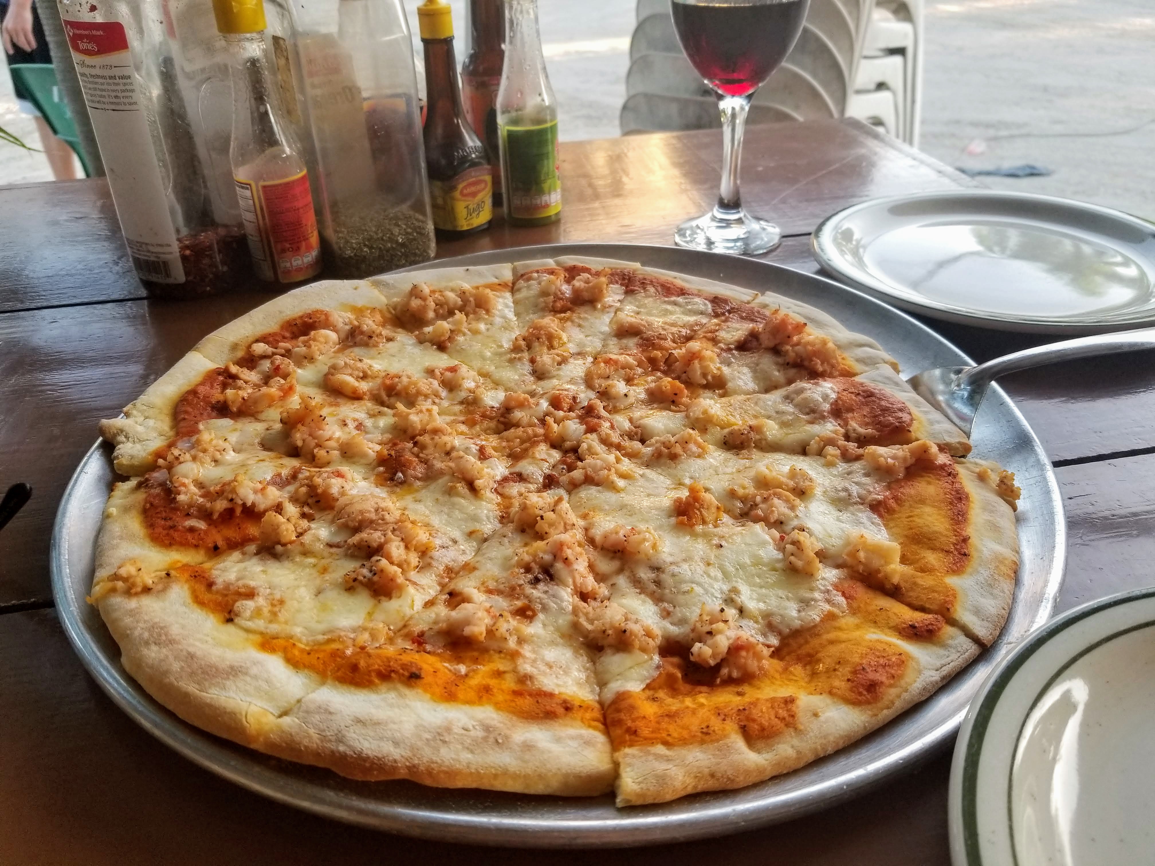 a large lobster pizza is in the foreground. in the background is a glass of red wine and an assortment of spices and hot sauces.